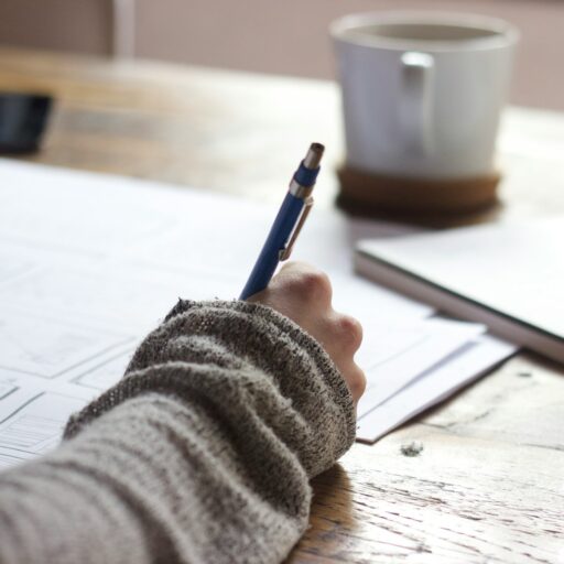 person writing on brown wooden table near white ceramic mug